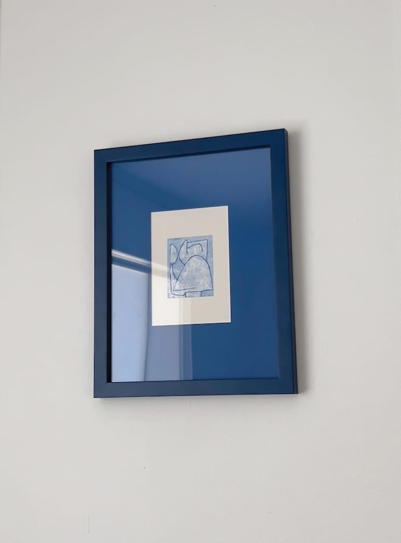 May include: A framed abstract art print. The artwork features a light blue design on a white background, set within a deep blue mat and frame. The piece is mounted on a white wall, creating a clean and modern aesthetic.