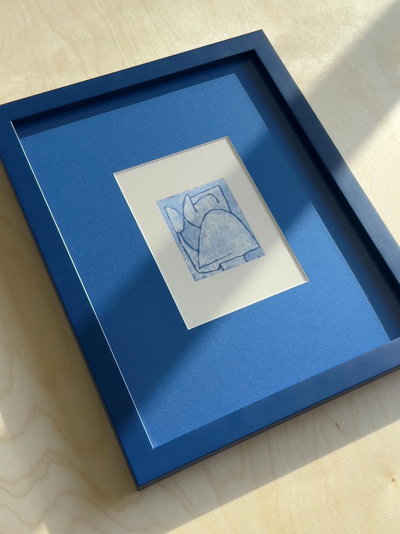 May include: A framed artwork featuring a blue abstract design on white paper. The artwork is set within a blue mat and a matching blue frame. The piece is displayed on a light wood surface.