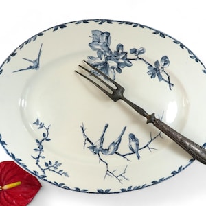Puede incluir: Plato ovalado blanco con diseño floral y de pájaros azules. Un tenedor plateado descansa sobre el plato. El borde tiene un patrón de hojas azules. Una flor de anturio roja está en la esquina inferior izquierda.
