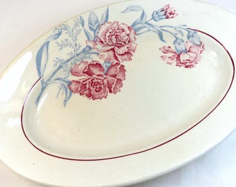 May include: An oval ceramic serving platter with an off-white base color. The platter features a floral pattern of pink carnations with light blue stems and leaves. A thin, dark red circular line accents the inner rim of the serving dish.