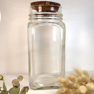 Peut inclure: Un bocal en verre clair et rectangulaire avec un bouchon en liège. Le bocal a une large ouverture et une forme légèrement incurvée. Le bouchon en liège est de couleur marron foncé. Le bocal est vide et posé sur une surface blanche.