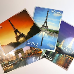 May include: A selection of postcards showcasing the Eiffel Tower and Parisian sights. Each card depicts a different view, including sunset, daytime reflections, and night scenes. The word "Paris" is printed on each postcard in a cursive font.