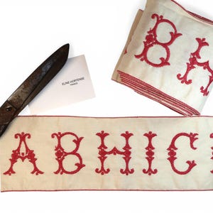 May include: Cream-colored fabric ribbons with embroidered red letters and decorative borders. The letters A, B, H, I, C, and D are visible. A pair of antique scissors and a small card are also present.