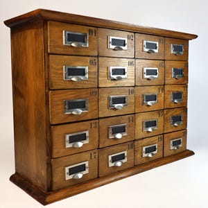May include: A wooden storage cabinet featuring twenty individual drawers arranged in a five-by-four grid. Each drawer is equipped with a silver metal label holder and a small pull handle, with black numbers printed on the wood above each drawer.