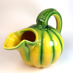 May include: A ceramic pitcher shaped like a pumpkin with a glossy finish. The body features alternating vertical stripes of bright yellow and vibrant green. It has a curved handle and a wide, scalloped pouring spout at the top of the vessel.