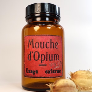Może przedstawiać: Bursztynowa szklana butelka apteczna z czarną zakrętką. Prostokątna czerwona etykieta z przodu zawiera czarny napis Mouche d'Opium, numer 0315 oraz Usage externe. Obok butelki znajdują się dwie wysuszone, jasnobrązowe torebki nasienne.
