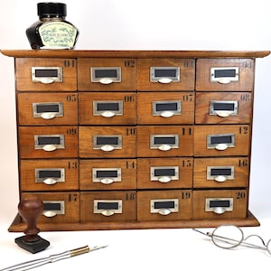 Può includere: Un mobiletto in legno con venti cassetti numerati, ciascuno dotato di porta etichetta e maniglia in metallo. Un flacone di inchiostro nero, un timbro in legno, un pennino in metallo e occhiali tondi sono disposti attorno al mobile.
