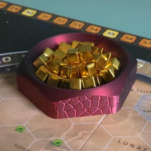 May include: A burgundy hexagonal bowl filled with gold game pieces, on a board game map. The bowl has a textured exterior. The game board features a map of Mars with labeled locations. The game pieces are gold-coloured.