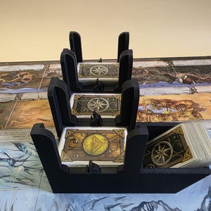 May include: Black card holders with a unique design, holding multiple decks of cards. The cards feature a compass rose design. The card holders are on a game board with a map design.