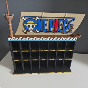 Può includere: Uno scaffale espositivo nero a più scomparti a forma di nave, dotato di una vela color sabbia con il logo blu di One Piece e un emblema a teschio. Lo scaffale presenta tre file di scomparti, ciascuno etichettato con testo di colore giallo.