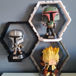 May include: Three hexagonal shelves with black and white frames display three pop culture figures. The top shelf has a green and red Boba Fett figure, the middle shelf has a silver Mandalorian figure, and the bottom shelf has a yellow Naruto figure.