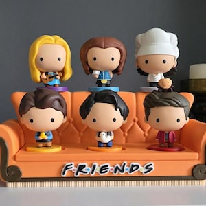 Può includere: Una collezione di sette figurine di personaggi dei cartoni animati della serie TV "Friends". Le figurine sono esposte su una base a forma di divano arancione con il logo dello show. I personaggi indossano vari abiti.