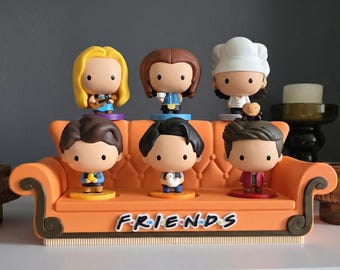 May include: A collection of seven cartoon figurines of characters from the TV show "Friends". The figurines are displayed on an orange couch-shaped base with the show's logo. The characters are in various outfits.