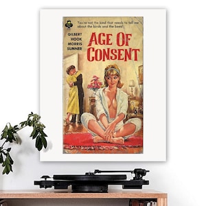 May include: A framed print of a vintage book cover titled "Age of Consent." The cover art features a woman in a white outfit, seated on a red rug, with a couple embracing in the background. Text includes the author's name and a quote.
