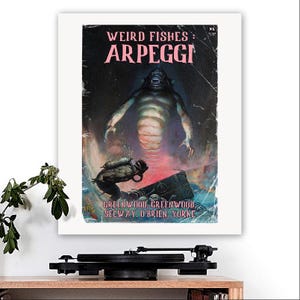 Könnte beinhalten: Ein gerahmter Druck eines Vintage-Horrorfilmplakats mit dem Titel "Weird Fishes: Arpeggi". Das Kunstwerk zeigt eine monströse Kreatur, die aus dem Wasser aufsteigt, mit einem Taucher im Vordergrund. Die Farbpalette des Posters umfasst Pink-, Blau- und Schwarztöne.