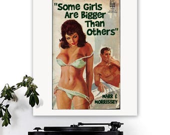 Op de afbeelding: Een ingelijste print van een vintage boekomslag met de tekst "Some Girls Are Bigger Than Others". De illustratie toont een vrouw in bikini en een man. De print wordt weergegeven op een houten oppervlak met een platenspeler.