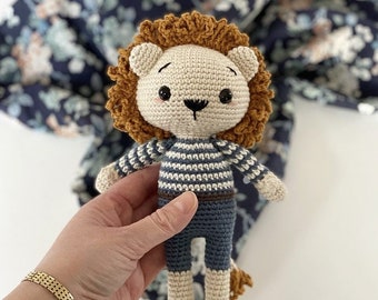 May include: A crocheted lion toy wearing a blue and white striped shirt and blue shorts. The lion has a brown mane and a cute face.
