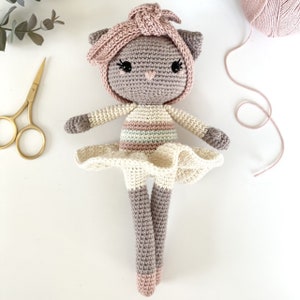 Könnte beinhalten: Eine gehäkelte Katzendolle, die ein rosa-weiß gestreiftes Kleid und eine rosa gehäkelte Mütze trägt. Die Puppe hat einen grauen Körper und rosa Wangen.