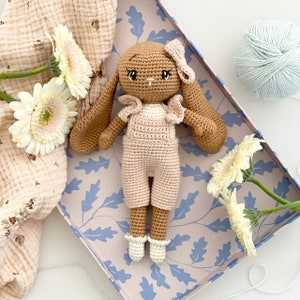May include: A crocheted bunny toy with brown fur, pink overalls, and a white bow. The bunny is sitting in a blue and white patterned box.