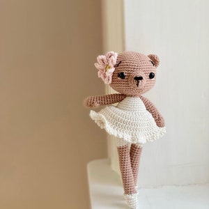 Può includere: Un orso di peluche marrone lavorato a uncinetto che indossa un tutù bianco e un fiore rosa sulla testa. L'orso è in piedi su una superficie bianca.