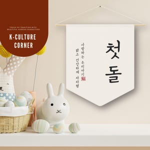 May include: A white banner with black Korean characters and text, hanging from a wooden rod. The banner reads "첫돌" and other Korean text. The banner is part of a K-Culture Corner display, with a white bunny figurine and decorative eggs.
