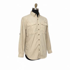 May include: A tan button-down shirt with two chest pockets and long sleeves. The shirt is displayed on a mannequin against a white background. The sleeves have a button to roll them up.