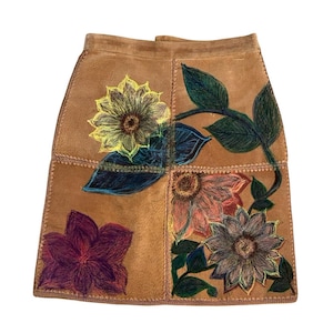 May include: A brown suede skirt with a patchwork design. The skirt features hand-drawn floral designs in yellow, pink, purple, and blue. The skirt is a mini-length and has a straight cut.
