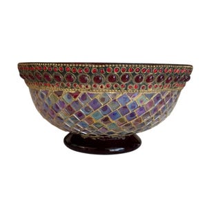 May include: A decorative bowl featuring a mosaic exterior of iridescent, multi-colored square tiles. The upper rim is adorned with a band of circular red stones set in gold-toned metal. The bowl rests on a solid, dark-colored circular pedestal base.