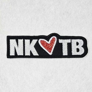 May include: A rectangular embroidered patch with a black background and a white border. The patch features the white embroidered letters NK and TB, separated by a red glittery heart shape in the center. The design is flat and stitched throughout.