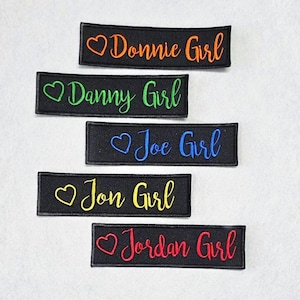 May include: Five rectangular black fabric patches with embroidered text and heart symbols. The patches feature the phrases Donnie Girl in orange, Danny Girl in green, Joe Girl in blue, Jon Girl in yellow, and Jordan Girl in red, all in script font.