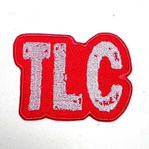 May include: A red embroidered patch featuring the letters "TLC" in white. The letters have a distressed, textured look, and the patch is a rounded rectangle.