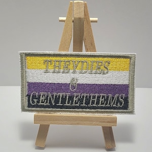 May include: A yellow, white, and purple embroidered patch with the text "Theydies & Gentlethems" on a small wooden easel.