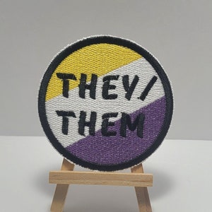 May include: A round, embroidered patch with a yellow, white, and purple design. The patch says "They/Them" in black text.