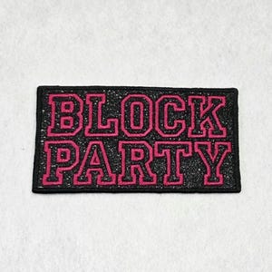 May include: A rectangular embroidered patch featuring the text BLOCK PARTY in bold, pink, varsity-style lettering. The background of the patch is a dark, glittery black material, and the entire design is framed by a solid black stitched border.