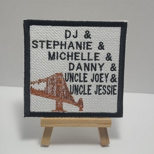 May include: A white fabric patch with black border and brown embroidery. The patch features a stylized image of the Golden Gate Bridge and the text "DJ & STEPHANIE & MICHELLE & DANNY & UNCLE JOEY & UNCLE JESSIE".