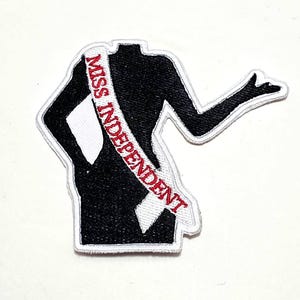 May include: A black patch in the shape of a silhouette of a person with a white sash. The sash reads "MISS INDEPENDENT" in red text. The patch has a white border.