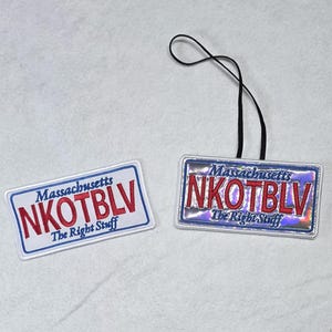May include: Two rectangular items resembling Massachusetts license plates. One is a white patch with red text reading NKOTBLV, Massachusetts, and The Right Stuff. The other is a holographic version of the same design with a black hanging loop.