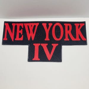 May include: A black patch with the words "NEW YORK IV" in red, stylized lettering. The patch has a rectangular shape with a smaller rectangular extension below the "NEW YORK" text. The background has a subtle woven texture.