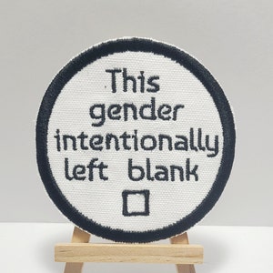Op de afbeelding: Witte ronde patch met zwarte borduurwerk die "Dit geslacht is met opzet leeg gelaten" zegt met een vierkant in het midden.
