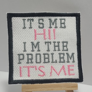 May include: White fabric patch with black border and pink and gray embroidery. The text reads "It's Me Hi I'm The Problem It's Me". The patch is displayed on a small wooden easel.