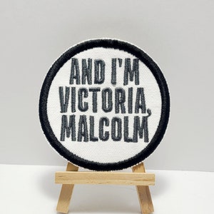 Może przedstawiać: Okrągła biała łatka z czarnym obramowaniem, z tekstem "AND I'M VICTORIA, MALCOLM" w czarnym hafcie. Łatka jest wyświetlana na małym drewnianym sztalugach.