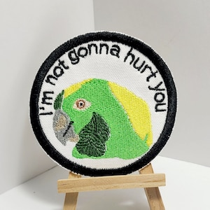May include: A round embroidered patch with a green parrot with yellow feathers and the text "I'm not gonna hurt you" in black.