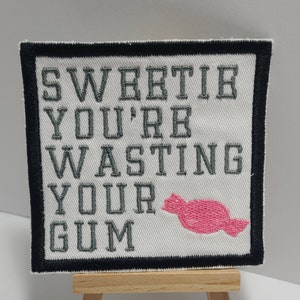 May include: A white fabric patch with black border, featuring the text "Sweetie you're wasting your gum" in gray embroidery. A pink gumdrop is embroidered to the right of the text.