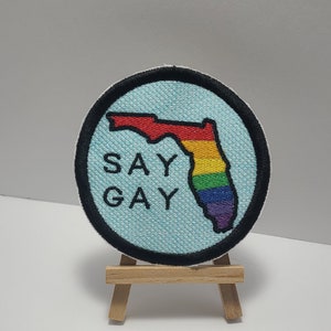 May include: Round patch with a light blue background and a black border. The patch features the outline of Florida in rainbow colors and the words "SAY GAY" in white. The patch is displayed on a small wooden easel.