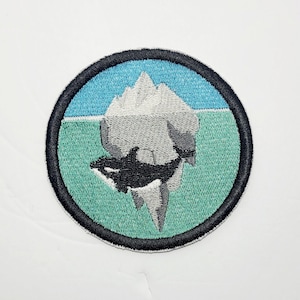 May include: Embroidered patch featuring an orca breaching beneath an iceberg. The design is set within a circular frame with a black border. The background shows a blue sky and green water. The orca is black and white, and the iceberg is gray.