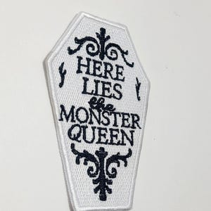 Puede incluir: Parche blanco en forma de ata&uacute;d con texto bordado en negro y adornos decorativos. El texto dice "HERE LIES the MONSTER QUEEN". El parche tiene un borde blanco y est&aacute; destinado a ropa o accesorios.