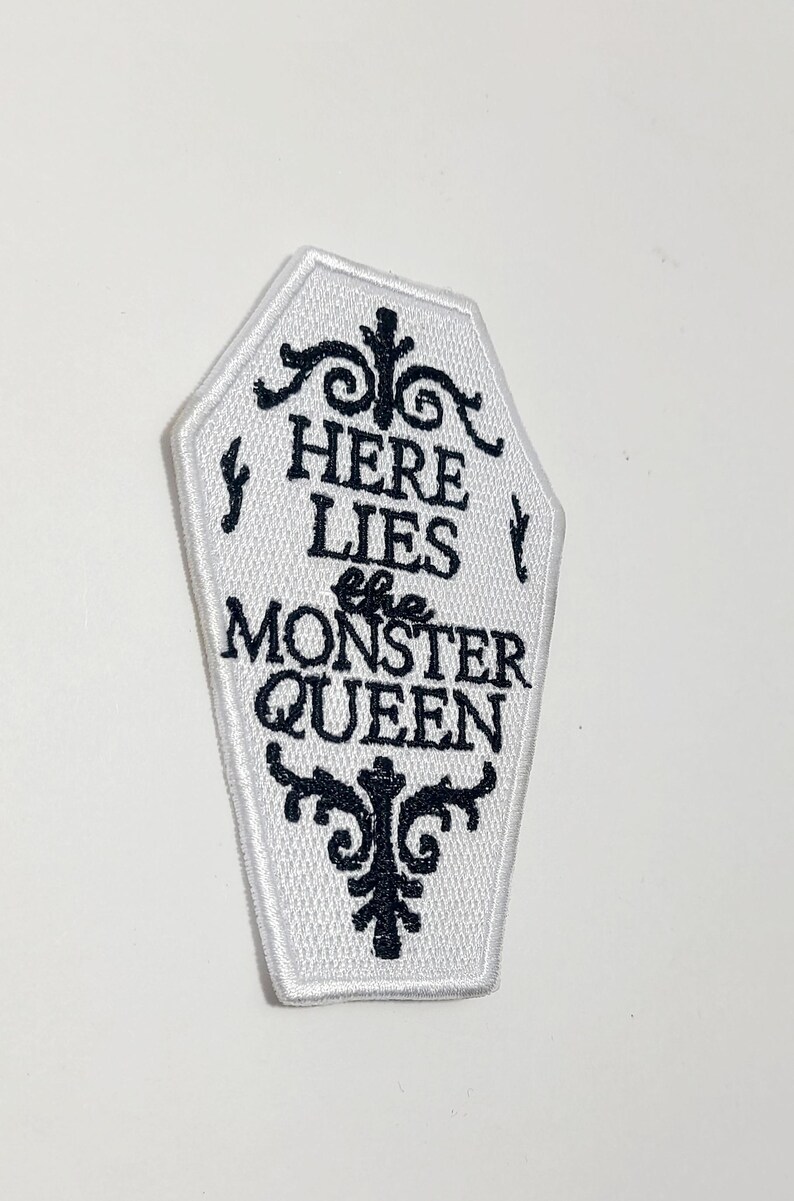 Puede incluir: Parche blanco en forma de ata&uacute;d con texto bordado en negro y adornos decorativos. El texto dice "HERE LIES the MONSTER QUEEN". El parche tiene un borde blanco y est&aacute; destinado a ropa o accesorios.