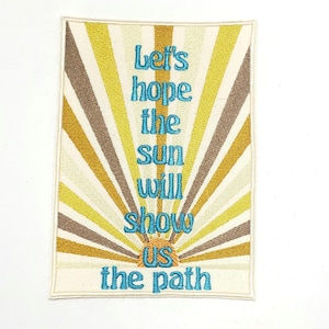 May include: Embroidered patch with a retro sunburst design in shades of yellow, brown, and cream. The patch features the text "Let's hope the sun will show us the path" in turquoise. The patch has a rectangular shape.