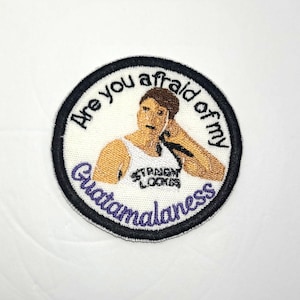 May include: A white round embroidered patch with black and purple stitching. The patch features a cartoon image of a man with the text "Are you afraid of my Guatemalaness" around the image.