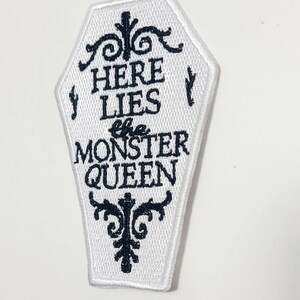 Puede incluir: Parche blanco en forma de ata&uacute;d con texto bordado en negro y adornos decorativos. El texto dice "HERE LIES the MONSTER QUEEN". Un parche de tem&aacute;tica g&oacute;tica y espeluznante para ropa o accesorios.